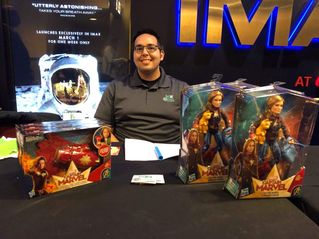 ReelOutreach's Jordan Maison sitting at a table in front of a theater with Captain Marvel toys laid out, waiting to check in attendees.